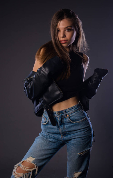 Portrait of an attractive trendy girl in denim ripped pants and leather jacket over black background - Fotó, kép