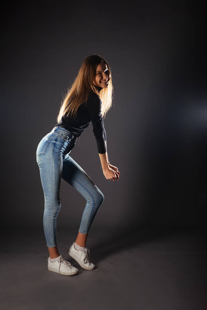 Side full-body portrait of a young female model in black top and jeans posing with one leg in front - Foto, Bild