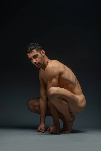 Portrait of a naked male model posing squatted on black studio background - Φωτογραφία, εικόνα
