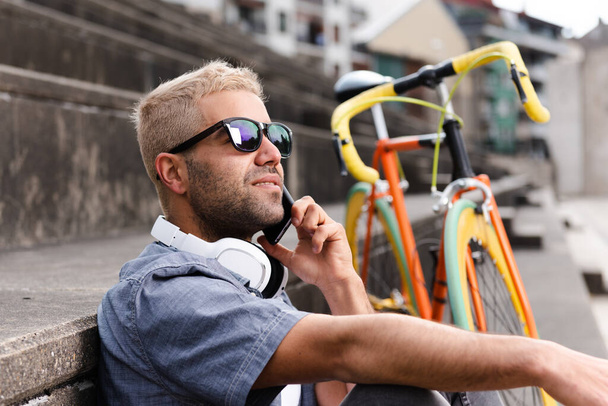 ヒスパニックフリーランサーともにブロンド髪の話彼の携帯電話の横にAヴィンテージ自転車. - 写真・画像