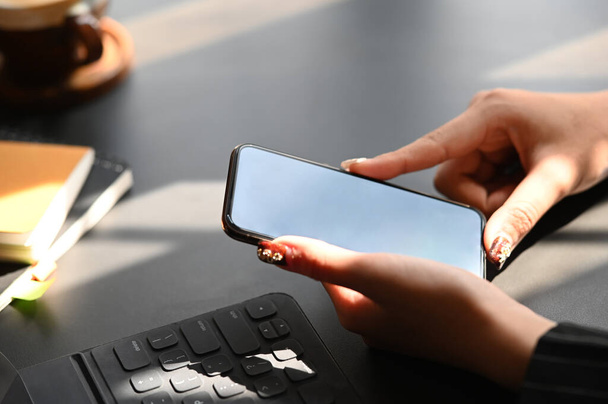 Ausschnitt aus der Hand einer Geschäftsfrau, während sie ihr Smartphone mit weißem Bildschirm auf dem modernen Arbeitstisch in der Nähe des Tablets mit Tastaturhülle, Notebook und Kaffeetasse hält. - Foto, Bild