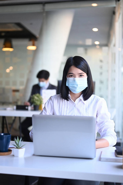 Portrait d'une femme d'affaires portant un masque de protection travaillant sur un ordinateur portable au bureau et de son collègue assis en arrière-plan. - Photo, image