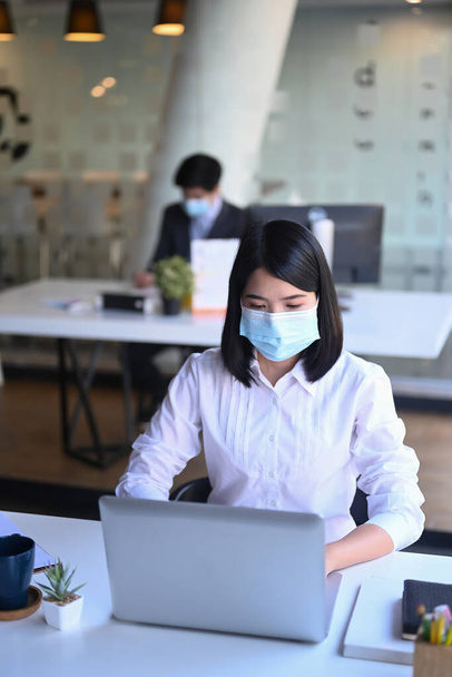 ノートパソコンで保護マスクを着けているビジネス女性と彼女の同僚はバックグラウンドで座っている. - 写真・画像