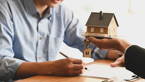 Cropped shot of House broker holding the sample house model in hand with his customer while writing on paper/ signing an agreement at the wooden table. Loan,Debt, selling/buying agreement concept. - Photo, Image