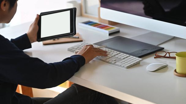 Καλλιεργημένη φωτογραφία του νεαρού στιλάτου παραγωγού που κρατά σε λευκό κενό tablet οθόνη και πληκτρολογώντας στο πληκτρολόγιο πάνω από το σύγχρονο φόντο γραφείο εργασίας. - Φωτογραφία, εικόνα