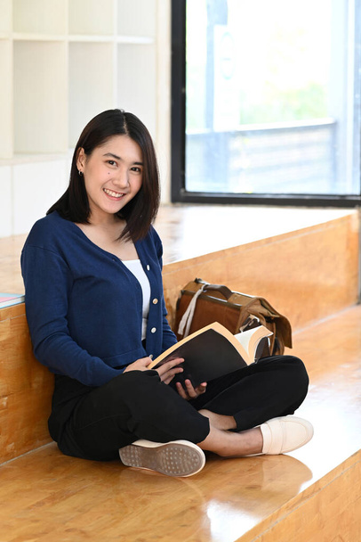 Portrét mladé asijské vysokoškolačky držící knihu a usmívající se před kamerou, zatímco sedí v knihovně. - Fotografie, Obrázek