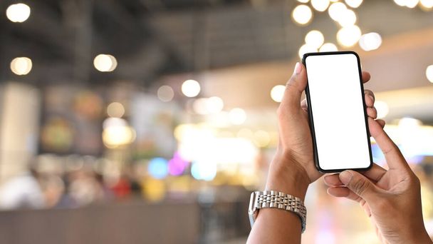 Imagen de cerca de la mano mientras sostiene el teléfono inteligente negro con pantalla blanca en blanco sobre fondo borroso del mercado nocturno. - Foto, imagen