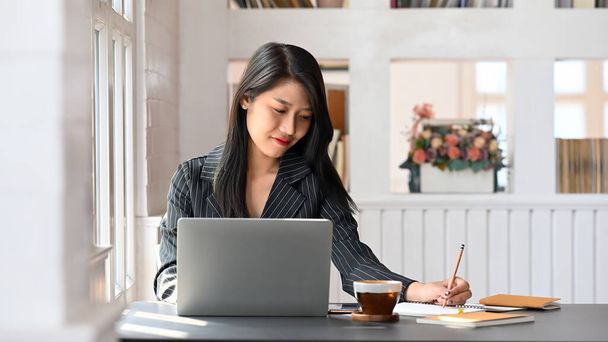 Νεαρή εκτελεστική γυναίκα γράφει στο σημειωματάριό της, ενώ κάθεται μπροστά από το φορητό υπολογιστή της στο σύγχρονο τραπέζι εργασίας με άνετο γραφείο ως φόντο. - Φωτογραφία, εικόνα