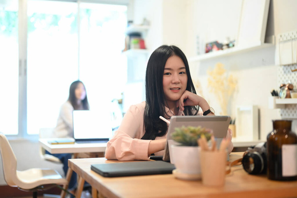 Sorrindo jovem designer gráfico feminino trabalhando em tablet digital enquanto se senta com seu colega no escritório. - Foto, Imagem