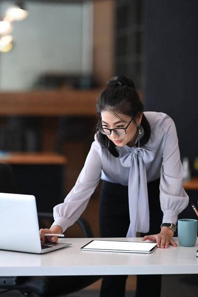 Portret bizneswoman w okularach stojącej i pracującej na laptopie w biurze. - Zdjęcie, obraz