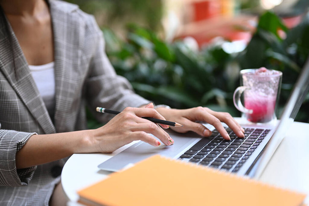Обрізаний знімок молодої жінки, що тримає ручку і друкує на клавіатурі ноутбука, сидячи в кафе
. - Фото, зображення