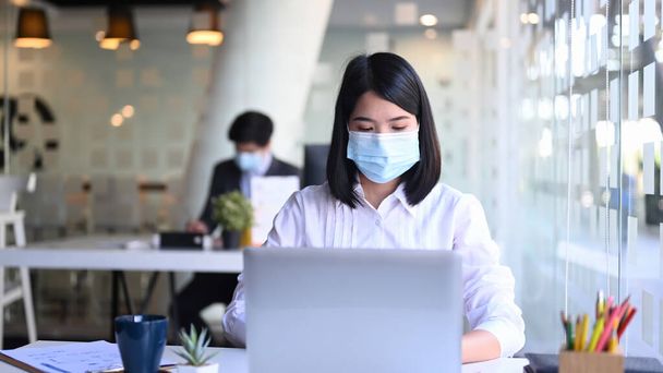 Bizneswoman w masce pracująca z laptopem i kolegami w tle w biurze. - Zdjęcie, obraz