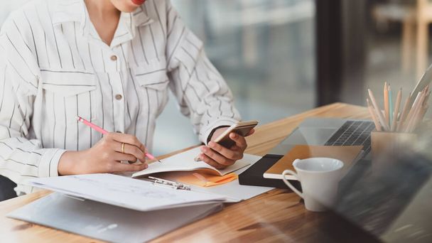 Oříznutý záběr mladé krásné ženy v pruhované košili držící smartphone v ruce a psaní / přičemž poznámka bílá sedí na moderním dřevěném stole s moderní kancelář jako pozadí. - Fotografie, Obrázek
