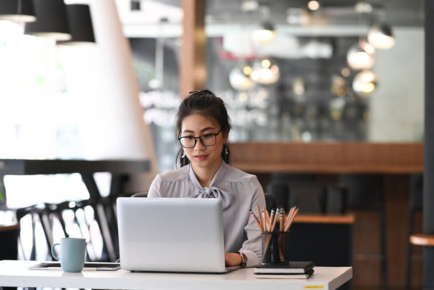 Young female employee in eyeglasses preparing financial marketing report on laptop at modern office. - Фото, зображення
