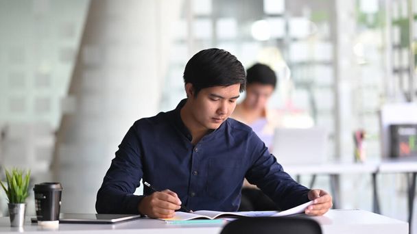 Фото молодого бизнесмена, пишущего на бизнес-документе у современного рабочего стола перед своим коллегой. - Фото, изображение