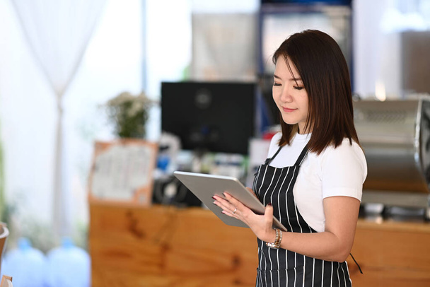 Jeune femme entrepreneur portant un tablier debout au café à la mode et en utilisant une tablette numérique. - Photo, image