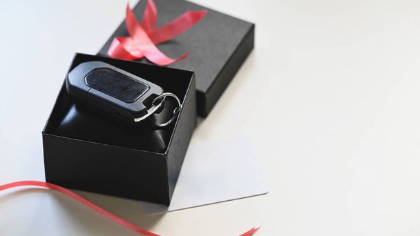 Közelkép a Digitális autó kulcs üzembe a fekete ajándék doboz piros szalaggal és kívánság kártya a fehér asztalon, mint a háttér. Meglepő Valentin napi ajándék koncepció. - Fotó, kép