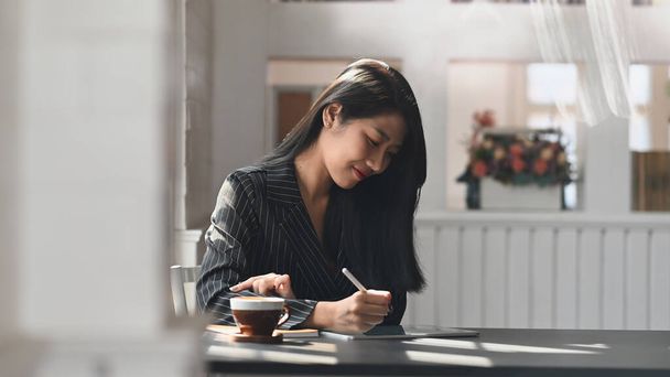 Foto de una joven ejecutiva en traje de rayas negras dibujando un plan de negocios por tableta de computadora en el moderno escritorio con una cómoda oficina como fondo. - Foto, imagen