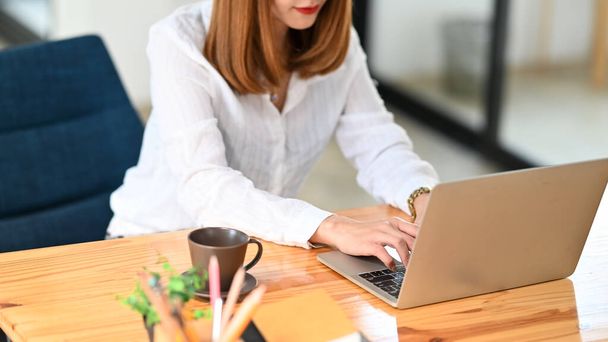 Foto einer jungen kreativen Frau, die als Sekretärin arbeitet, tippt auf einem Computer-Laptop mit weißem leeren Bildschirm am hölzernen Arbeitstisch mit geordnetem Büro als Hintergrund. - Foto, Bild