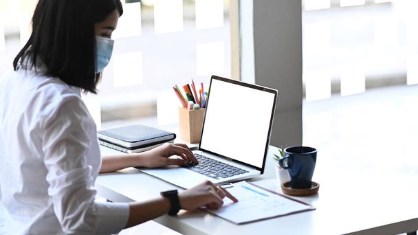 Вид збоку бізнес-леді в захисній масці, що працює на ноутбуці та аналізує діловий звіт на білому столі
. - Фото, зображення