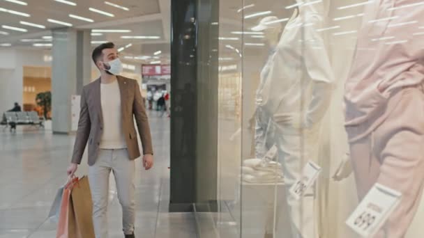 Tiro medio del hombre caucásico joven en ropa elegante y máscara facial caminando a lo largo de las pantallas de la ventana de tiendas de ropa de moda en un gran centro comercial moderno que lleva un montón de bolsas de compras - Metraje, vídeo