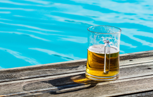 Plain pint glass of cold lager beer sitting on edge of blue swimming pool - Fotoğraf, Görsel