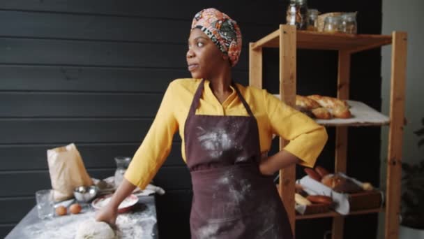 Zoom in shot of beautiful afro-american female αρτοποιός με ποδιά στέκεται με το χέρι στο σωρό στην κουζίνα, κοιτάζοντας κάμερα και χαμογελώντας - Πλάνα, βίντεο