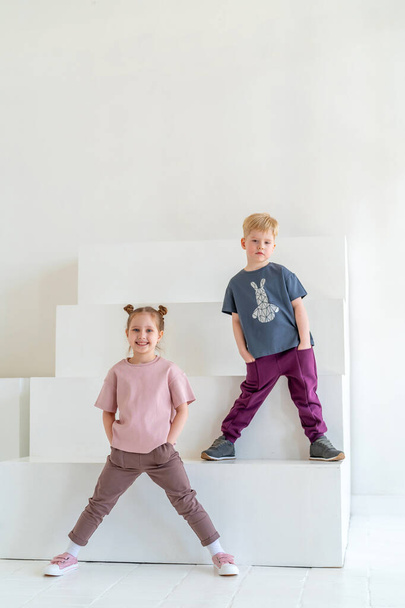 Little happy boy and girl in fashionable clothes smile, pose on background white wall studio. the concept of advertising children's clothing, emotional children, dynamic image. Copy space - Photo, Image