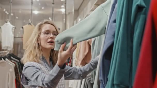 Vita-up shot di bella giovane donna in occhiali alla moda shopping per i vestiti in negozio moderno smistamento felpe appese su rotaie - Filmati, video