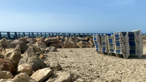 Montón de tumbonas en la playa de arena en el fondo de un mar no muy tormentoso y cuyas olas se rompen en el rompeolas. HD - Metraje, vídeo