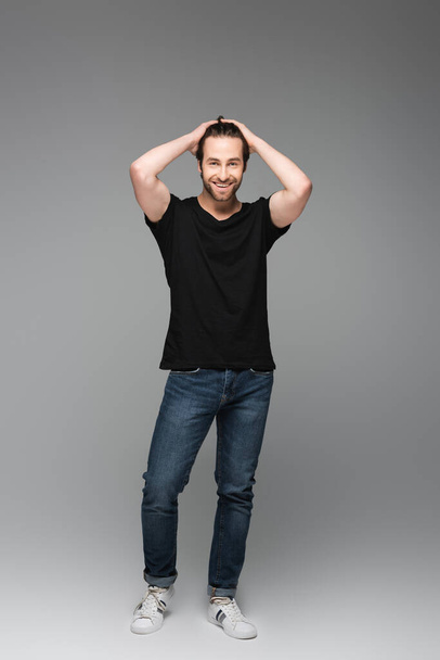 full length of bearded man in jeans and black t-shirt posing and smiling on grey - Foto, Imagem