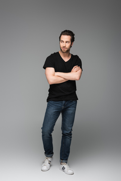 full length of bearded man in jeans and black t-shirt posing with crossed arms on grey - Foto, Imagem