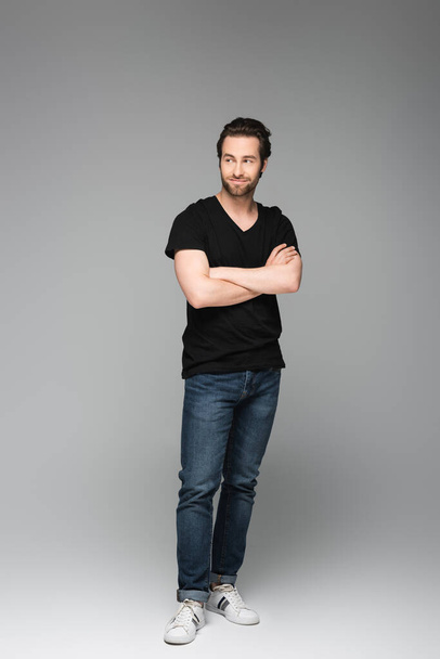 full length of good-looking man in jeans and black t-shirt posing with crossed arms on grey - Photo, image