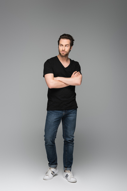 hombre barbudo serio en jeans y camiseta negra posando con los brazos cruzados sobre gris - Foto, imagen