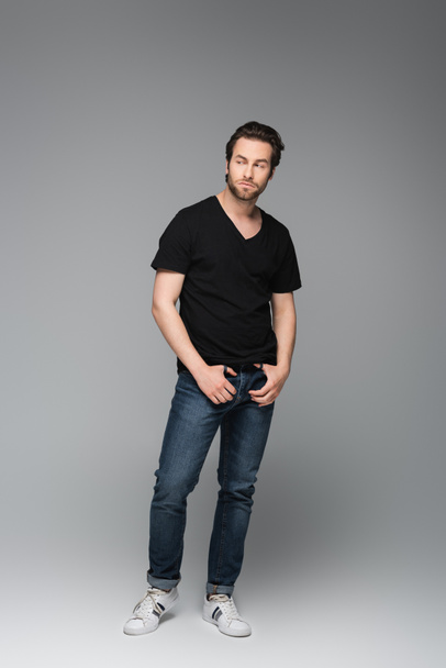 full length of bearded man in jeans and black t-shirt posing while looking away on grey - Fotografie, Obrázek