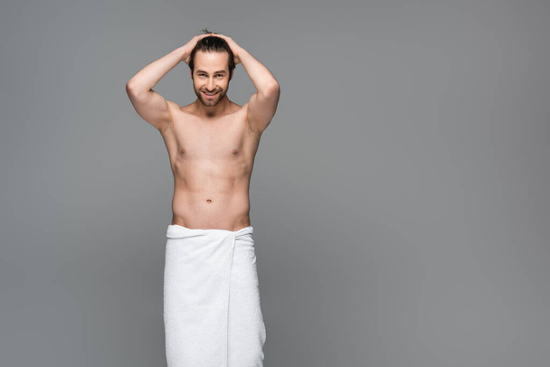 happy muscular man wrapped in towel adjusting hair isolated on grey - Фото, изображение