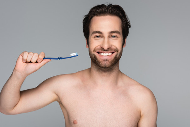 Neşeli ve gömleksiz bir adam diş fırçasını gri renkte tutuyor. - Fotoğraf, Görsel