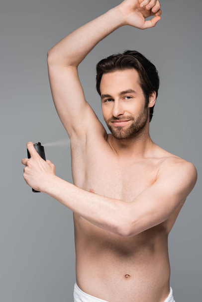 smiling and shirtless man with raised hand spraying antiperspirant isolated on grey - Zdjęcie, obraz