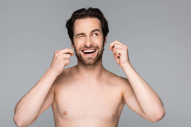 shirtless man winking eye while flossing teeth isolated on grey - Foto, afbeelding