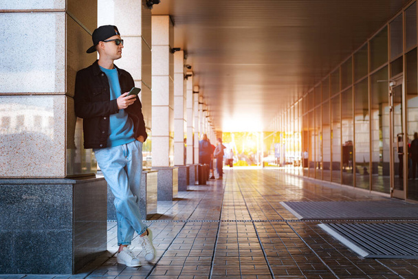 trendy stijlvolle man zwarte cap zonnebril met smartphone hoofdtelefoon achtergrond stedelijke zonsondergang - Foto, afbeelding