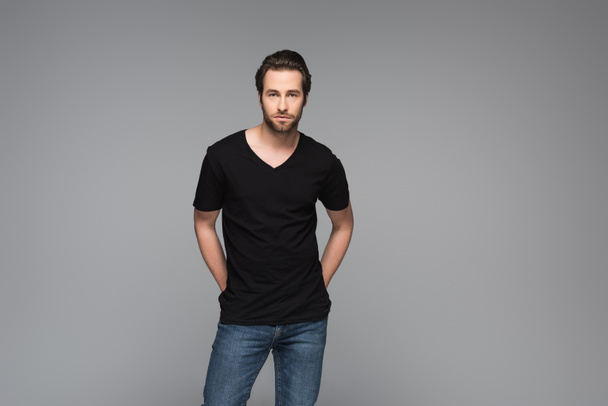 bearded man in black t-shirt and jeans posing with hands behind back isolated on grey - Photo, image