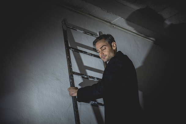 Ein junger erwachsener Mann steht mit geschlossenen Augen, meditiert oder denkt nach, während er, auf einer Leiter sitzend, nach Spanien greift. Er trägt lässig einen schwarzen Mantel. - Foto, Bild