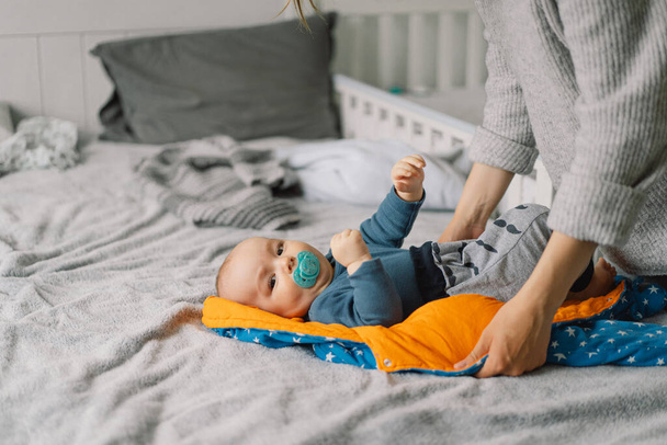 Mom dresses cute little baby boy in a jumpsuit. Happy young mother playing with baby on bed. Happy motherhood. Infant baby. Mother's day - Photo, Image