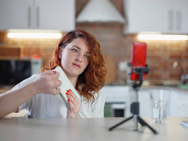 Τεχνολογία και blogging έννοια. Redhair γυναίκα καταγραφή βίντεο concent για το blog της στο σπίτι και κοιτάζοντας smartphone στο τρίποδο στο τραπέζι της κουζίνας, streaming online podcast στο κινητό τηλέφωνο. - Φωτογραφία, εικόνα