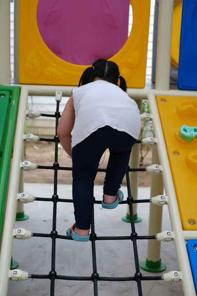 Una vista trasera de una linda chica asiática gordita joven subiendo por una rampa en un gran gimnasio de la selva al aire libre colorido, haciendo su camino a la parte superior de la estructura. - Foto, imagen