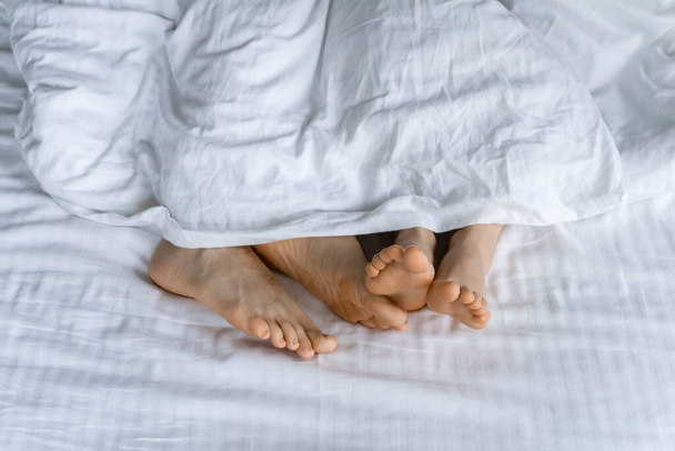 Goedemorgen en wakker worden. Voeten onder de dekens in bed in de ochtend. Womens voeten onder de dekens in een wit bed. Relax, slaap, rust concept. - Foto, afbeelding