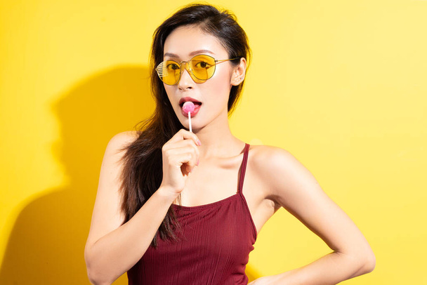 Beautiful asian woman in red swimsuit and eating lollipops on a yellow background - Фото, зображення