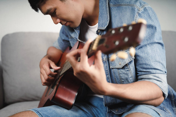 Насолоджуйтесь красивим азіатським чоловіком, який практикує або грає на гітарі на дивані у вітальні
  - Фото, зображення