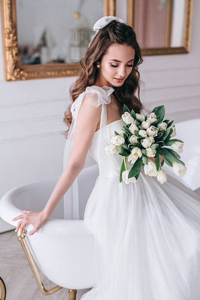Beautiful bride with a wedding bouquet sitting on sofa indoors in white studio interior like at home. - Fotó, kép