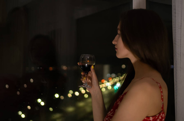 Молодая темноволосая женщина в красном платье с ремнями и бокалом красного вина стоит у окна и смотрит на ночную жизнь города. Селективный фокус - Фото, изображение